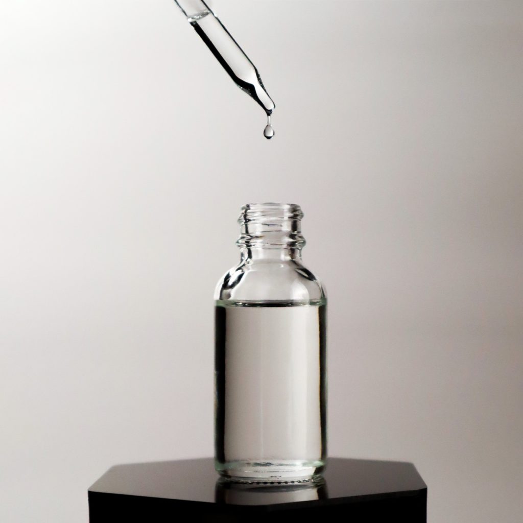 Clear cosmetic fluid in open dropper bottle on black hexagonal podium. Dropper dropping fluid into open bottle