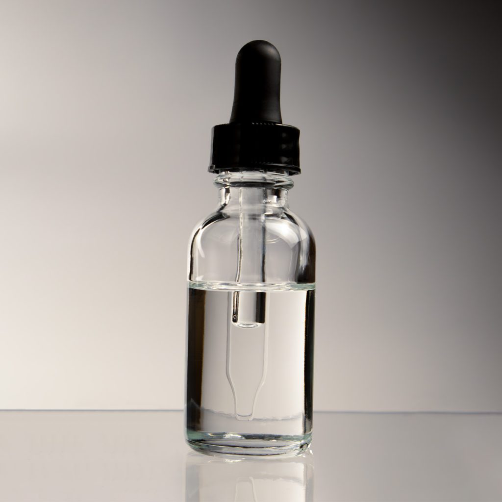 Clear cosmetic fluid in closed dropper bottle on reflective surface. White/grey background