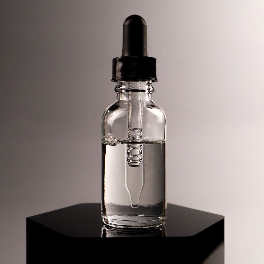 Closed clear cosmetic fluid in dropper bottle, air bubbles in dropper, on black hexagonal podium
